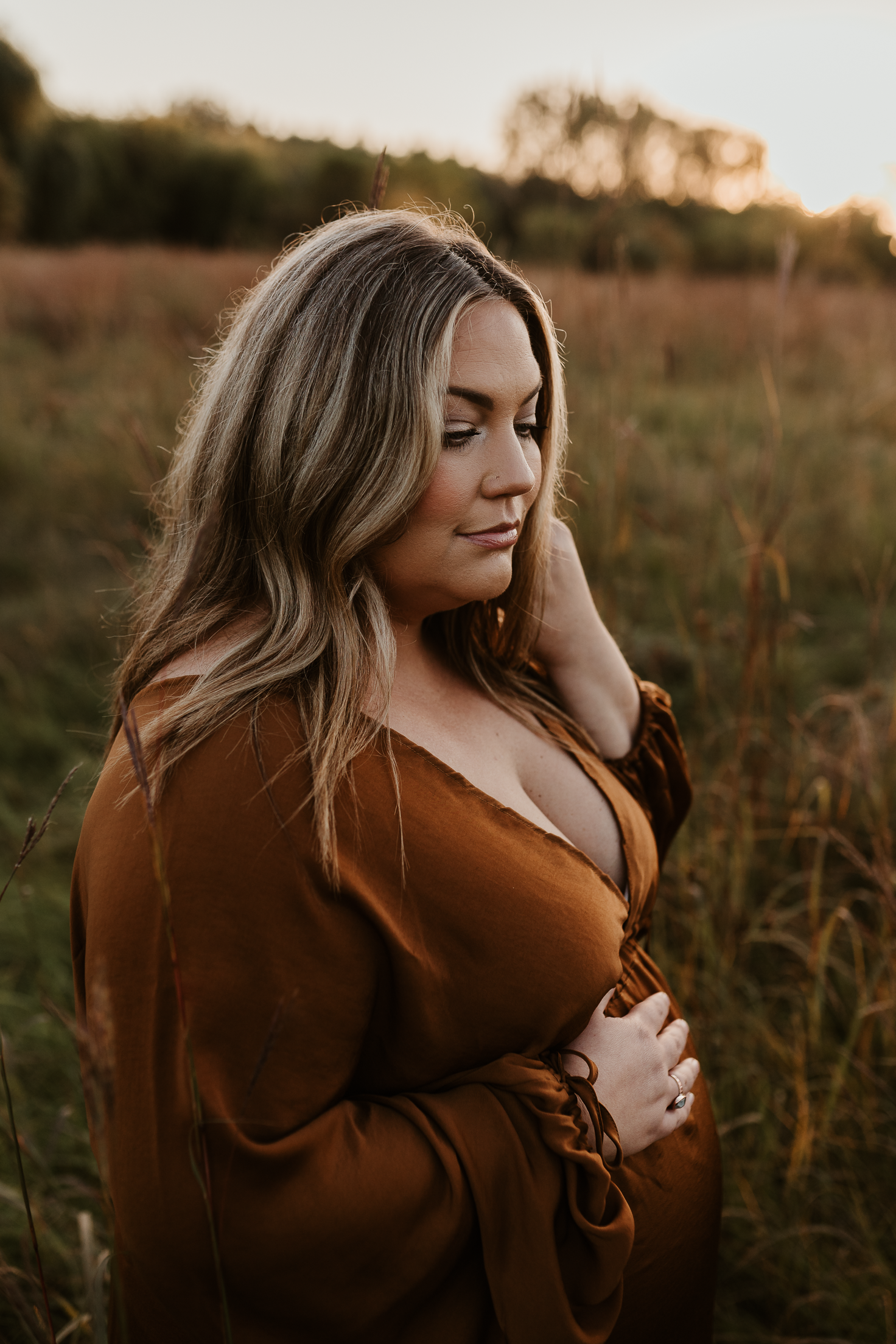 Pregnant woman in a golden bronze dress holding her belly in a field during sunset.